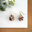 A pair of beaded, neutral-toned earrings rest on a white board with a cork texture in the background and a succulent leaf in the top left.