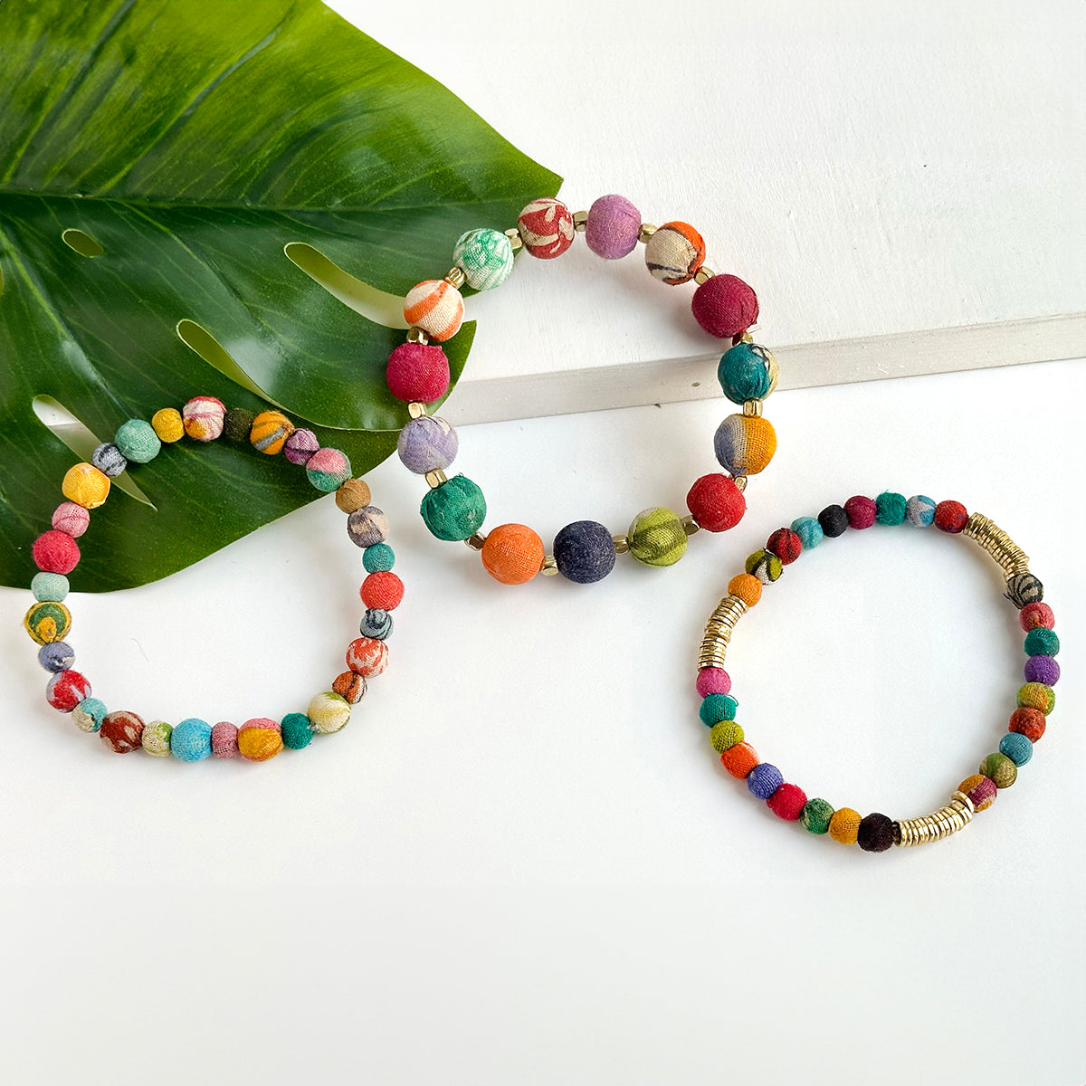 Three multicolor, beaded bracelets rest on a white backdrop with a large green leaf in the corner.