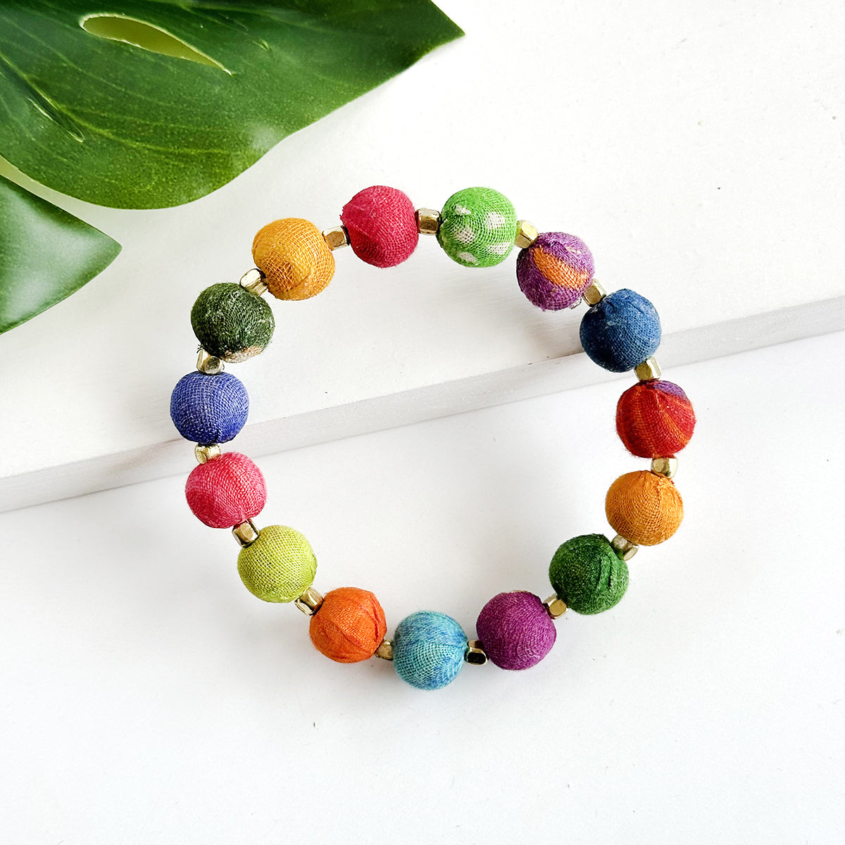 A multicolor, beaded bracelet rests against a white background with a green leaf.