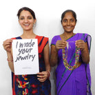 Two woman in Saris smile. One holds a beaded necklace and the other holds a paper sign that reads "I made your jewelry."