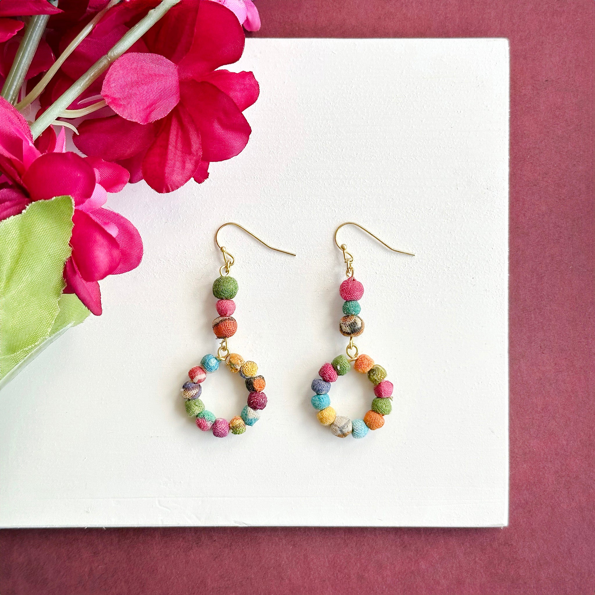 A pair of two earrings rests on a white surface with a dark pink flower is in the left corner.