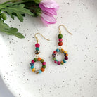 A pair of beaded, multicolor earrings rest on a speckled white surface with a pink and green flower rests in the top left.