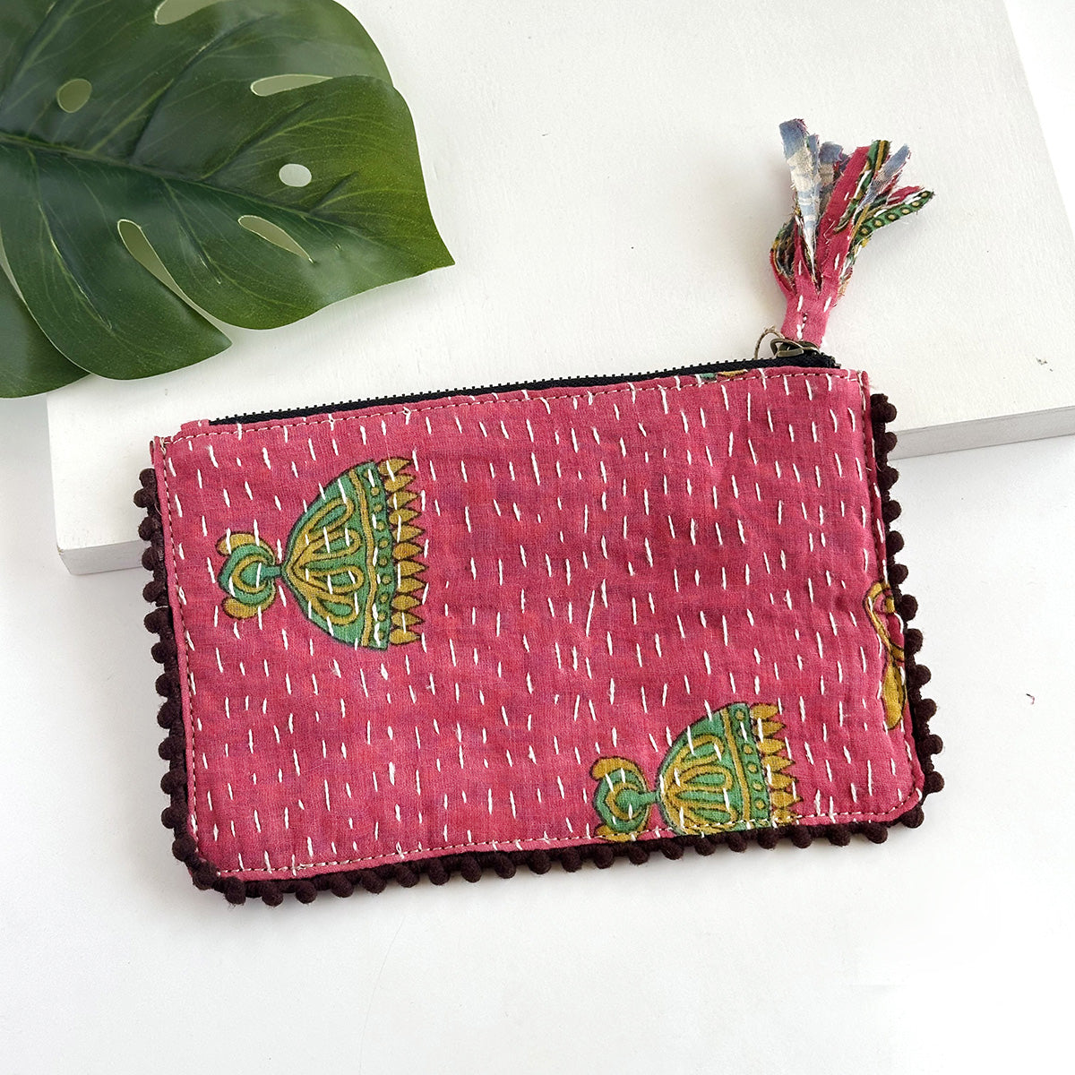 A pink rectangular pouch against a white background with a large leaf in the background.