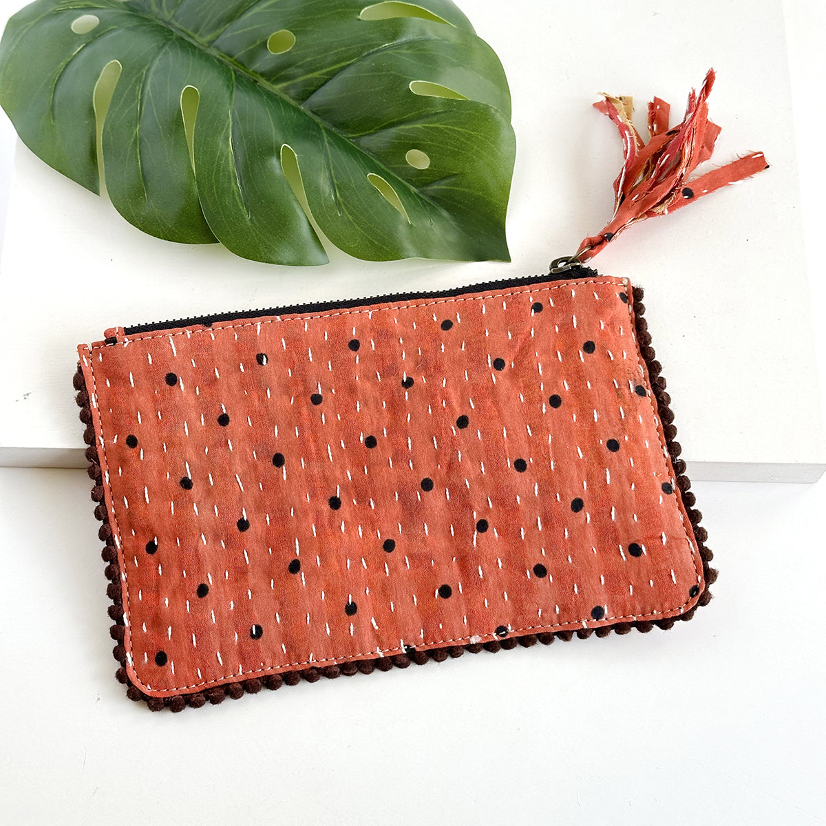 A polkadotted orange rectangular pouch against a white background with a large leaf in the background.