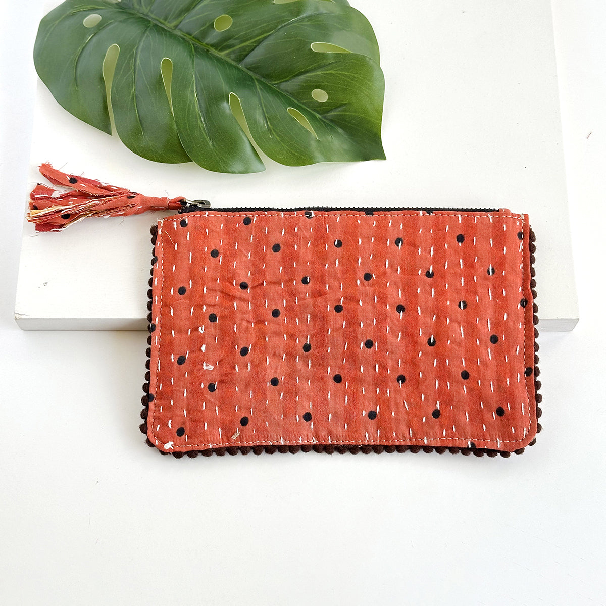 A polkadotted orange rectangular pouch against a white background with a large leaf in the background.