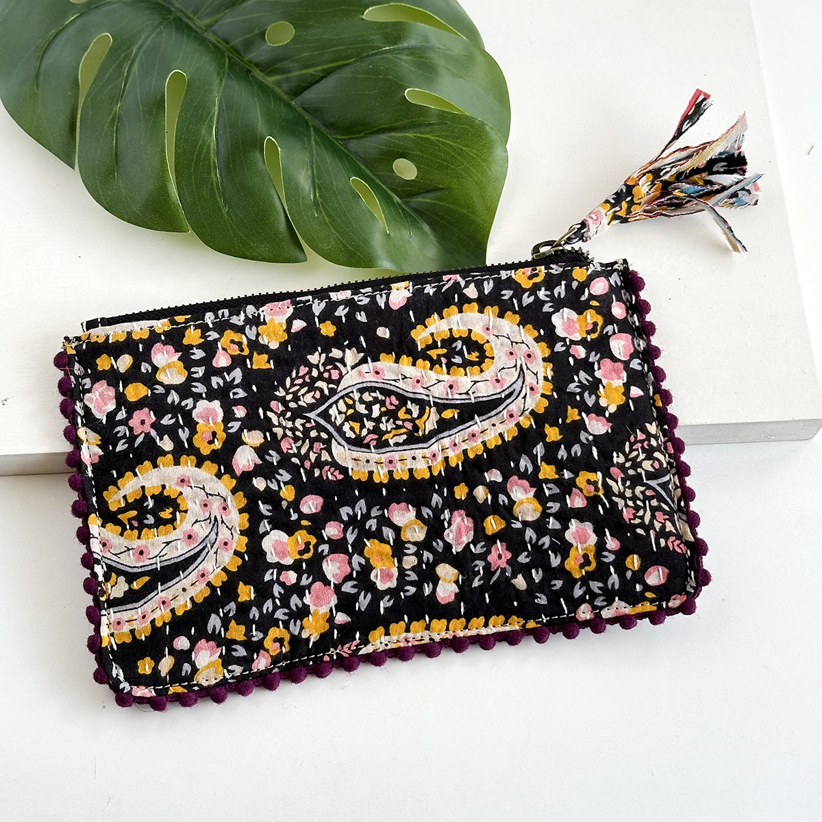 A black floral-patterned rectangular pouch against a white background with a large leaf in the background.