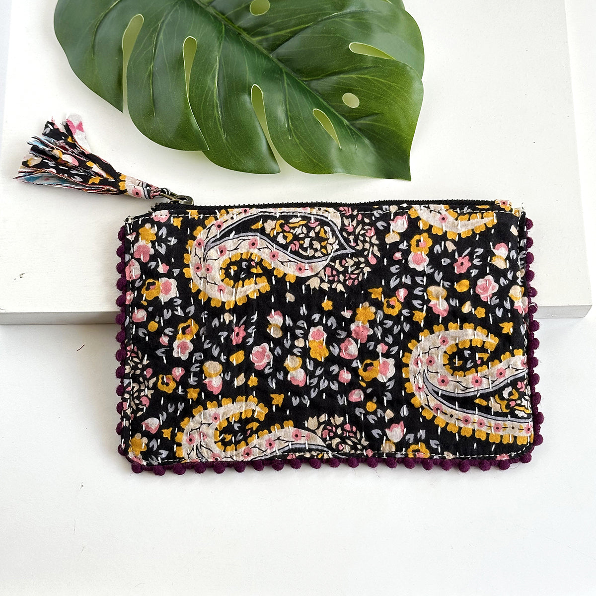 A black floral-patterned rectangular pouch against a white background with a large leaf in the background.