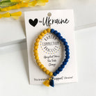 A blue and yellow beaded bracelet tied to a card that reads "Love to Ukraine" against a white background with a yellow flower to the left.