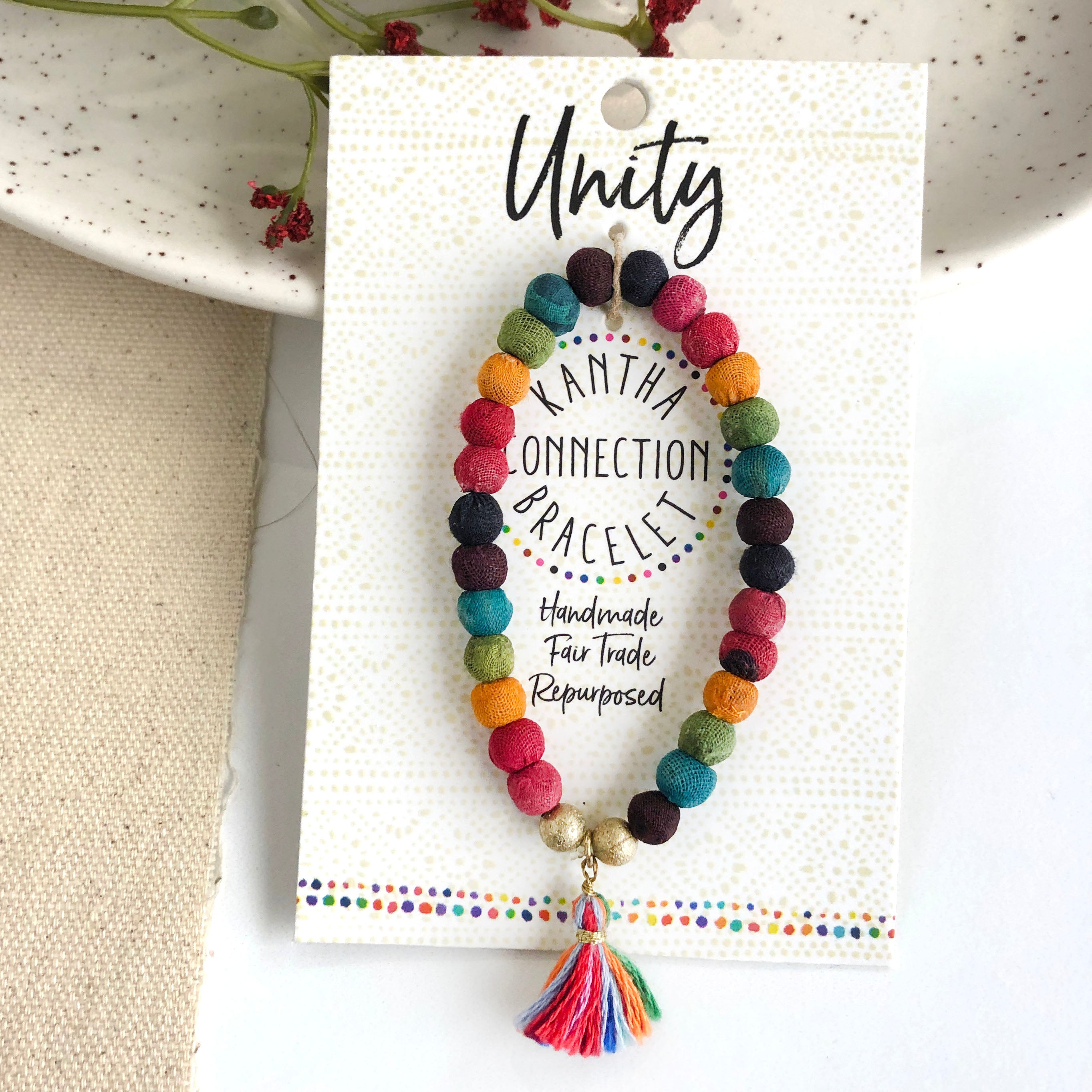 A rainbow beaded bracelet tied to a card that reads "Unity" against a white background with a green leaf to the left.