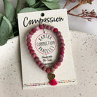 A pink beaded bracelet tied to a card that reads "Compassion" against a white background with a green leaf to the left.