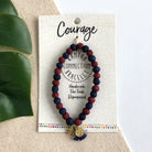 A red and blue beaded bracelet tied to a card that reads "Courage" against a white background with a green leaf to the left.