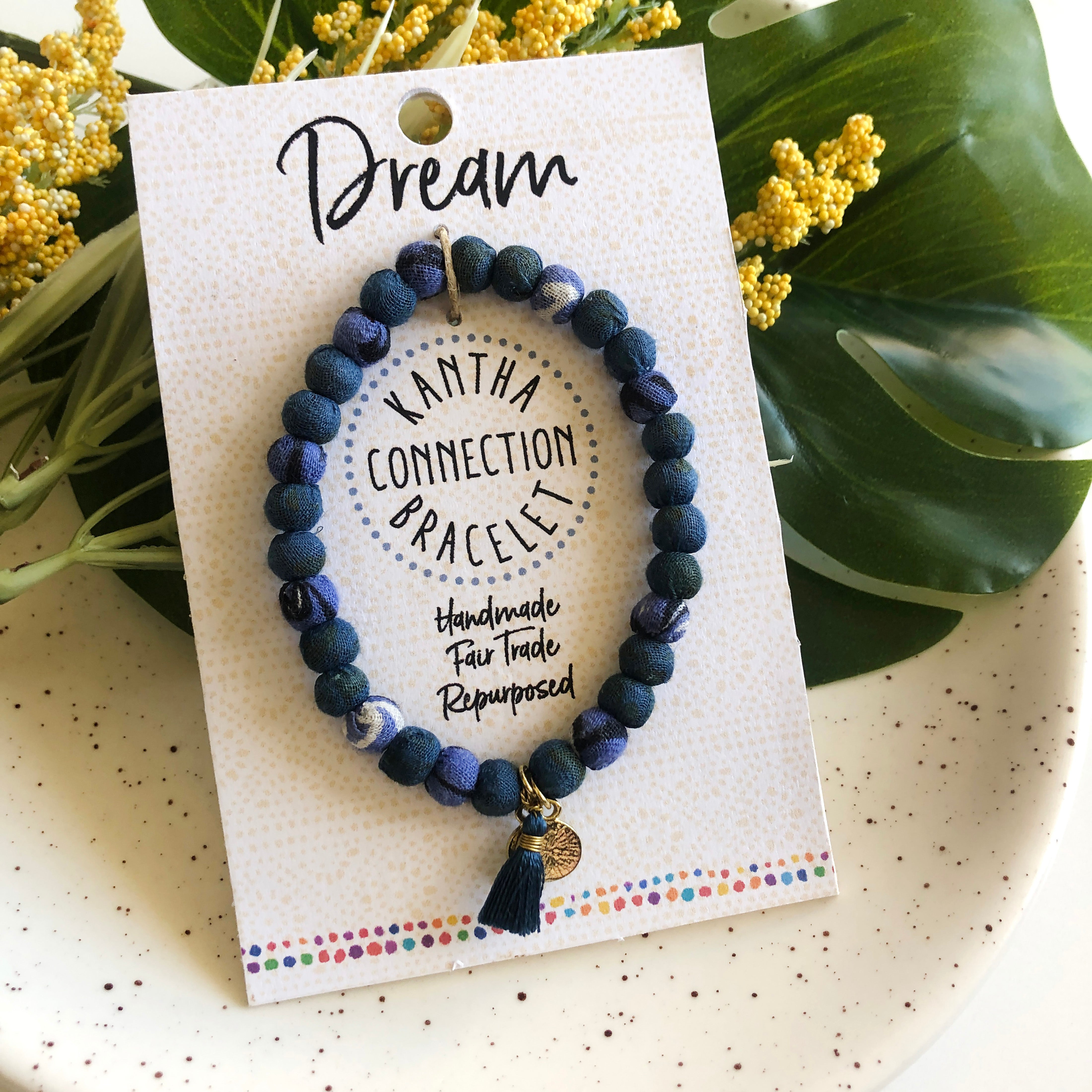 A blue beaded bracelet tied to a card that reads "Dream" against a white background with a green leaf to the left.