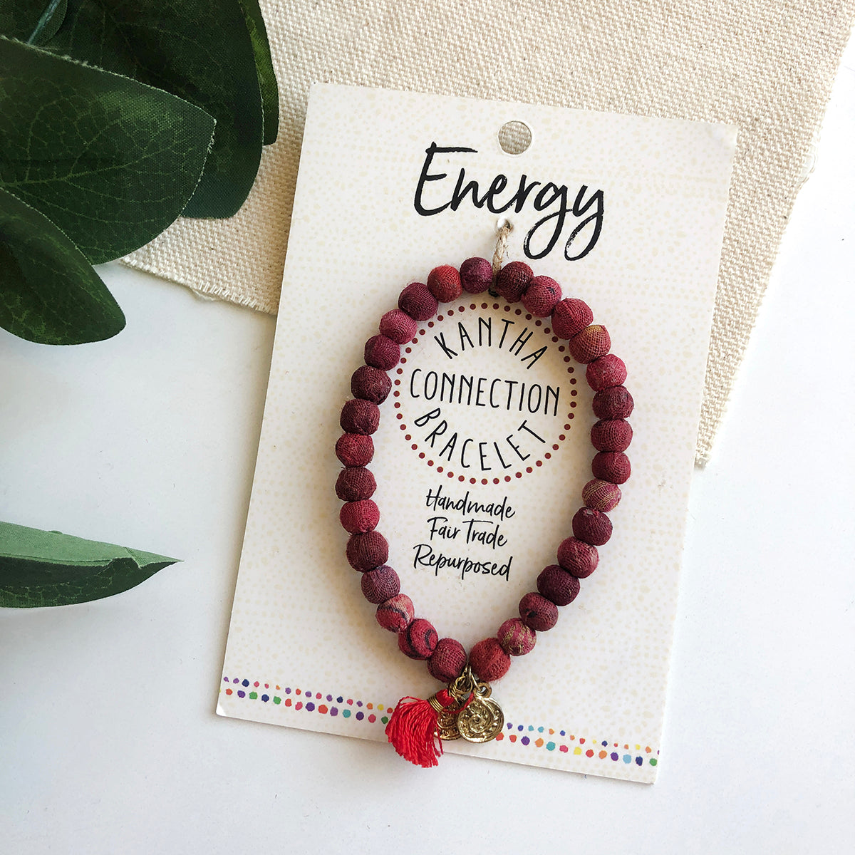 A red beaded bracelet tied to a card that reads "Energy" against a white background with a green leaf to the left.