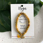 A yellow beaded bracelet tied to a card that reads "Hope" against a white background with a green leaf to the left.