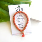 An orange beaded bracelet tied to a card that reads "Joy" against a white background with a green leaf to the left.