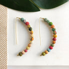 A pair of multicolor, arc-shaped earrings rests on a white surface with a green leaf in the background.