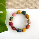 A colorful beaded bracelet against a white background with a tan woven fabric at the top of the image and a green leaf on the left.