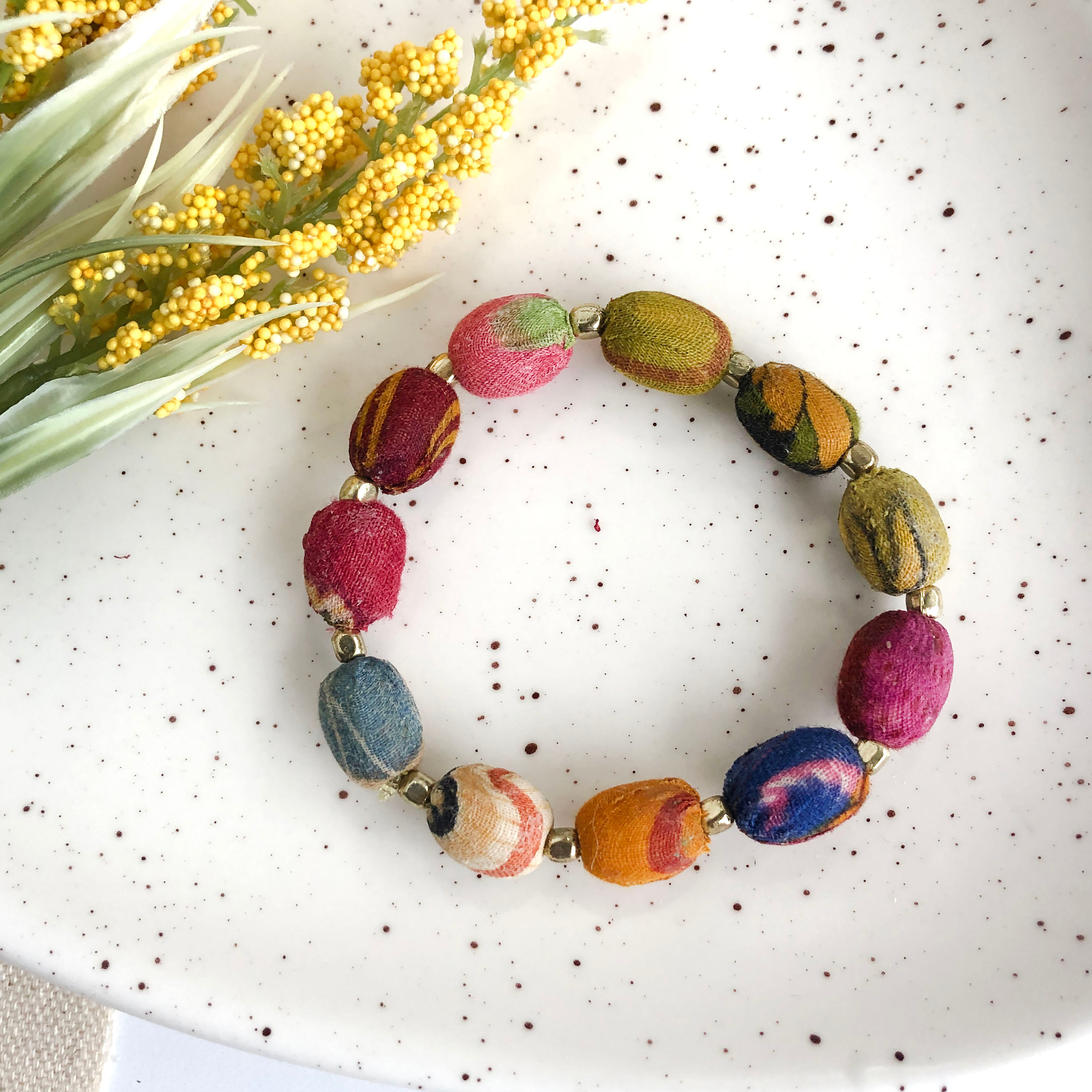 A colorful, beaded bracelet rests on a white speckled surface with a yellow flower in the corner.