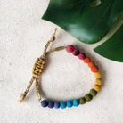 A beaded bracelet in rainbow-hues rests against a linen-surface with a green leaf.