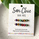 Two beaded multicolor bobby pins are attached to a packaging card and rest on a green leaf.