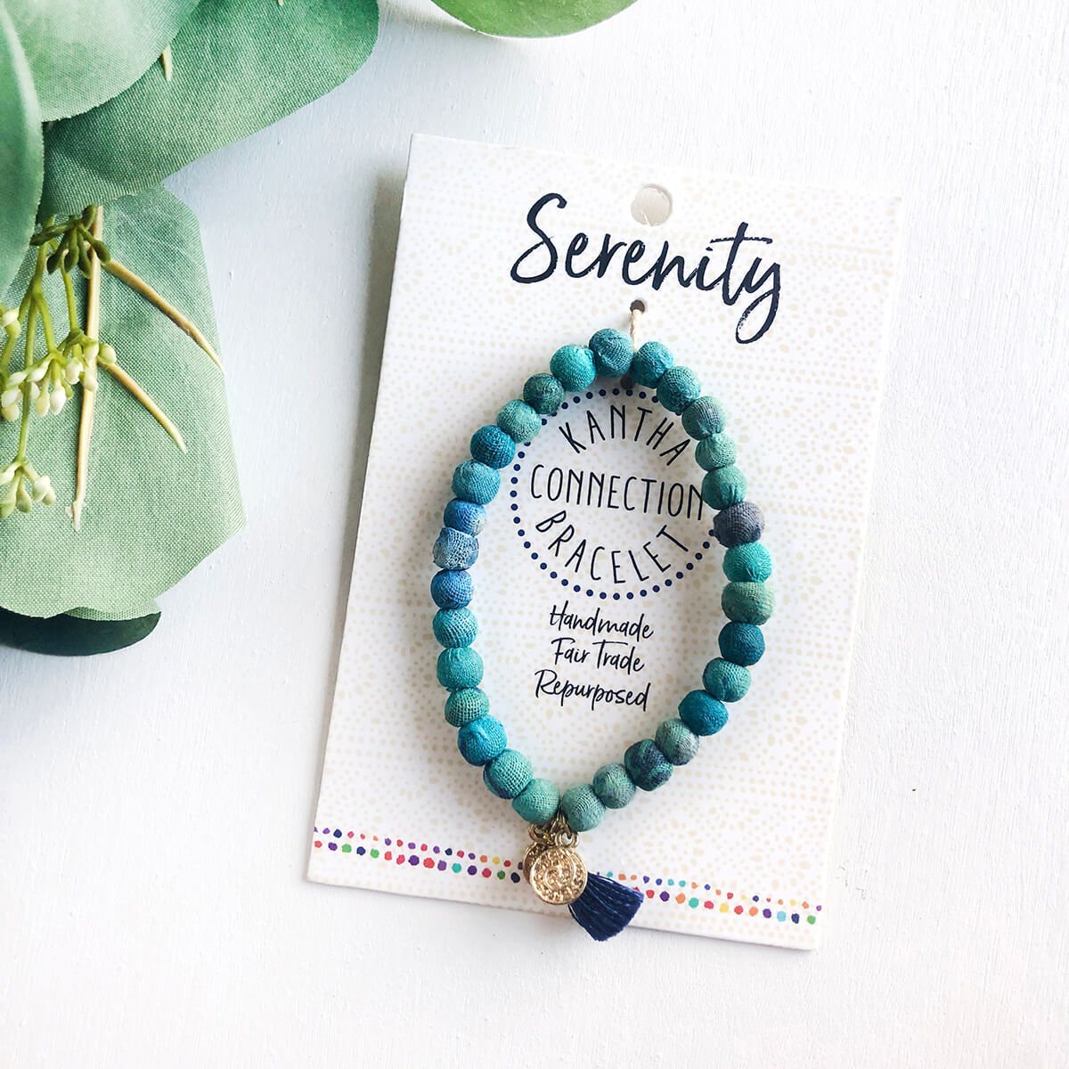 A blue beaded bracelet tied to a card that reads "Serenity" against a white background with a green leaf to the left.