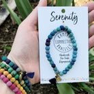 A woman holds a carded blue bracelet in her hand, while showing the rainbow of bracelets on her wrist.