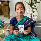 A woman in a blue Sari smiles while holding a carded blue bracelet that reads "Serenity".