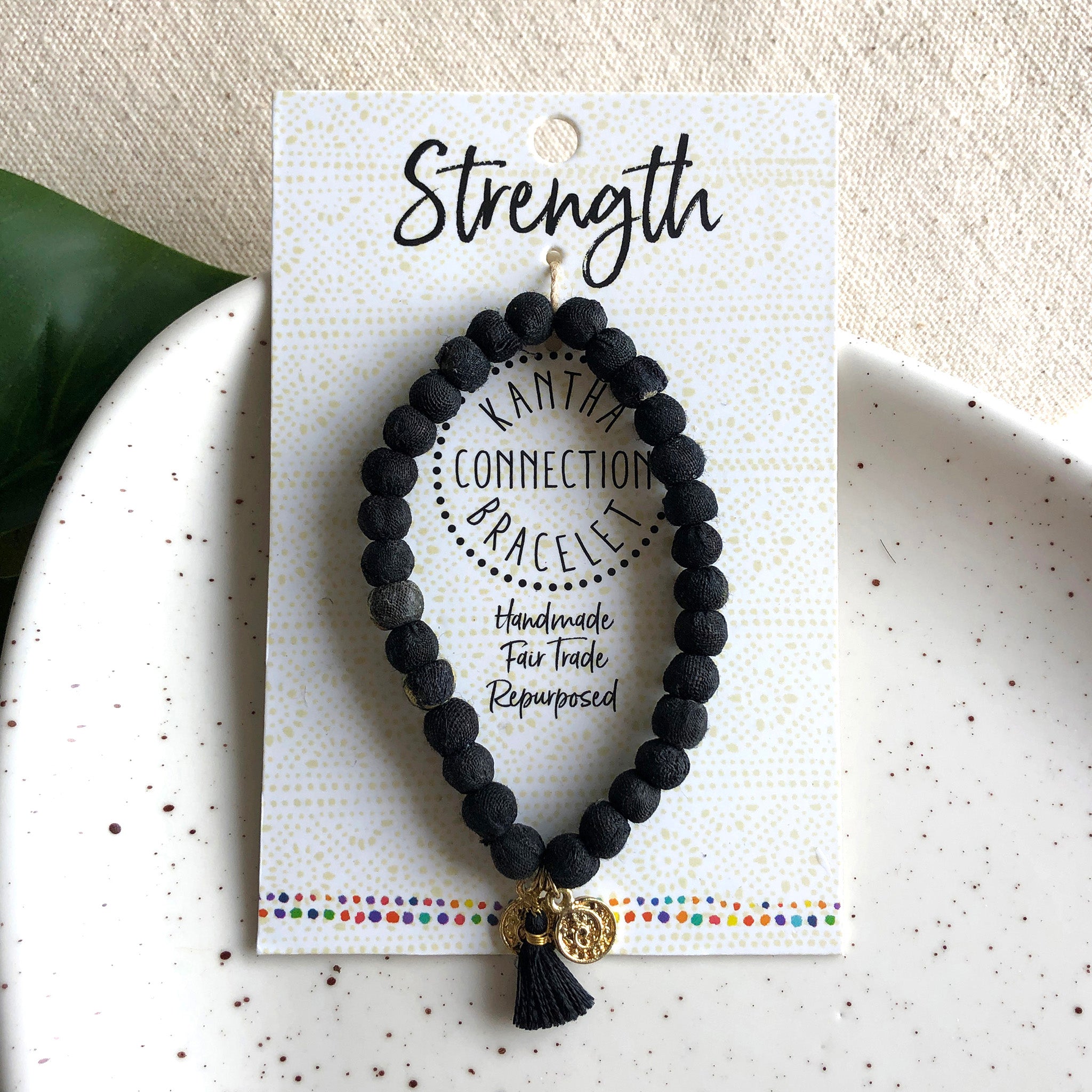 A black beaded bracelet tied to a card that reads "Strength" against a white background with a green leaf to the left.