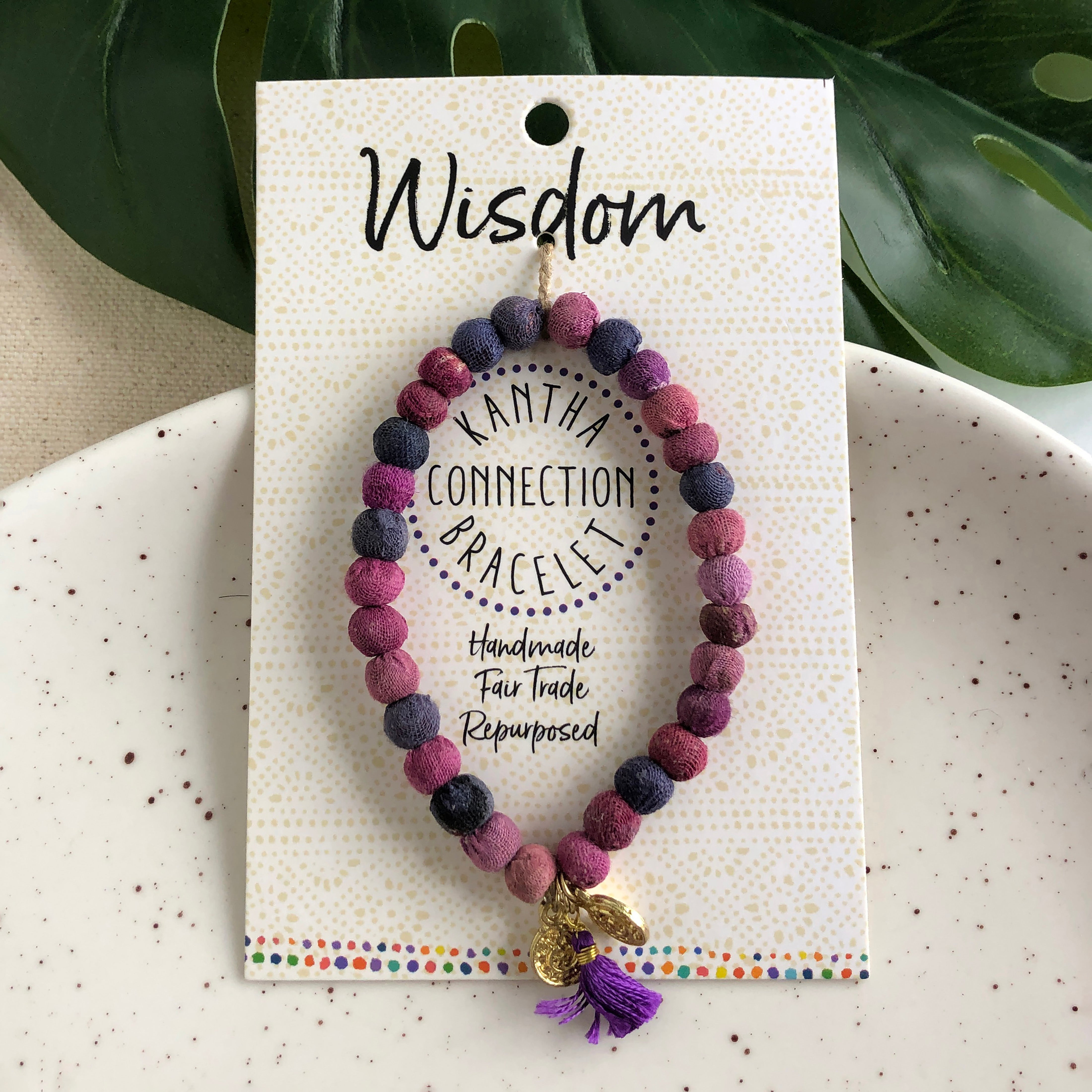 A purple beaded bracelet tied to a card that reads "Wisdom" against a white background with a green leaf to the left.